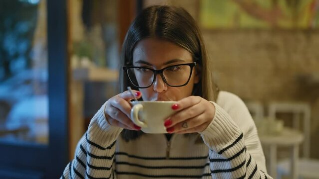 A young woman drinks coffee in a cozy cafe, portraying relaxed indoor leisure with her elegant glasses and manicured nails