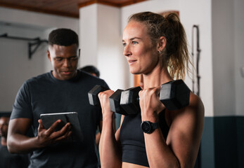 Woman lifts dumbbells in gym as man watches tablet - obrazy, fototapety, plakaty