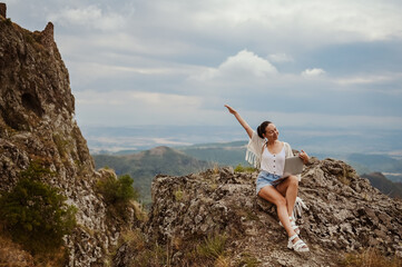 Young woman freelancer traveler working online anywhere outdoors using laptop enjoying mountain peak view. Happy female downshifter holding computer in unusual amazing view workplace at summer