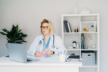 Friendly female middle aged doctor shows pills organizer to patient on laptop in clinic office. Video call online, treatment remote, pharmacy, supplement advice, medical healthcare. Telemedicine