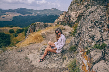 Naklejka na ściany i meble Young woman freelancer traveler working online anywhere outdoors using laptop enjoying mountain peak view. Happy female downshifter in sunglasses holding computer in unusual workplace at summer