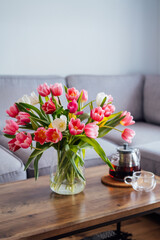 Focus on vase of fresh tulips bouquet and just brewed tea pot and cup on coffee table with gray couch sofa in modern leaving room. Cozy, comfortable relax at home. Time to take a break. Vertical card.