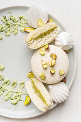 Top view of sweet macaron or french macaroon meringue-based confection dessert with pistachio flavour buttercream decorated with crushed nuts and cream cheese served on plate on white wooden table