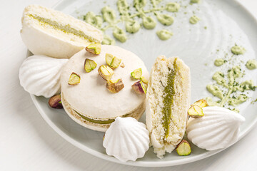 Close view of sweet crunchy macaron or french macaroon meringue-based confection with pistachio flavour ganache decorated with crushed nuts and cream cheese served on plate on white wooden table