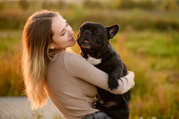 Young Caucasian woman holds and hugs dog in park during walk at sunset. Beloved dog is in arms of owner. Concept of pet care, love for pet, affection.