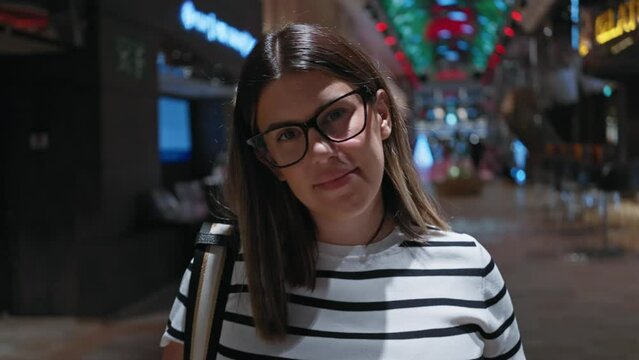 A smiling woman with glasses, in a striped shirt, poses inside a luxurious cruise ship with colorful lighting in the background.