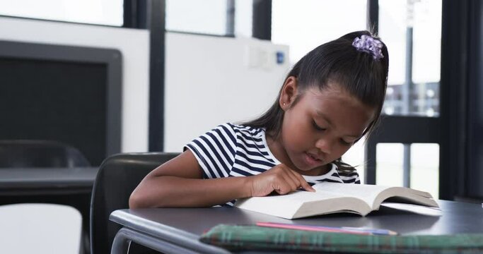 In a school classroom, a biracial young girl is focused on reading a book