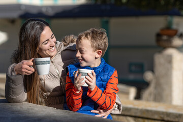 A mother shares hot chocolate with her young son on a picnic area, while they look each other smiling