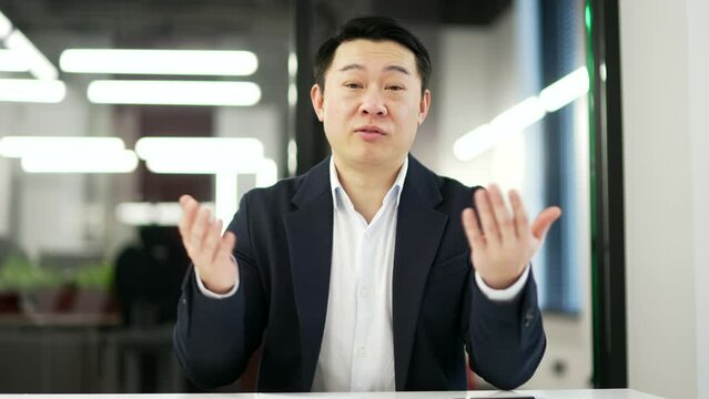 Webcam view. Asian businessman talking on a video call looking at the camera sitting in office. A man in a formal suit has a business meeting, speaks remotely, communicates at an online conference