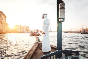 Fototapeten Arabic man with traditional clothes on the top of the boat, on the dubai river © oneinchpunch