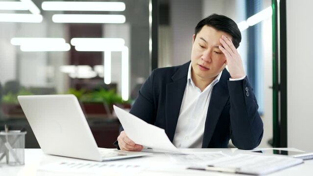 Overworked asian businessman is tired doing paperwork sitting at workplace in business office. A mature man in formal suit is overtired and bored. Overload exhausted financier has difficulties at work