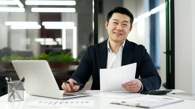 Portrait of a happy asian accountant in a formal suit sitting at desk at workplace in business office. Head shot of a smiling mature financier doing paperwork, working on laptop and looking at camera