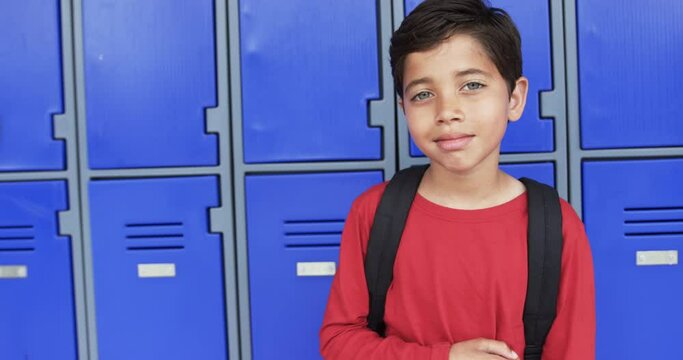 In a school hallway, a young biracial student stands before blue lockers with copy space