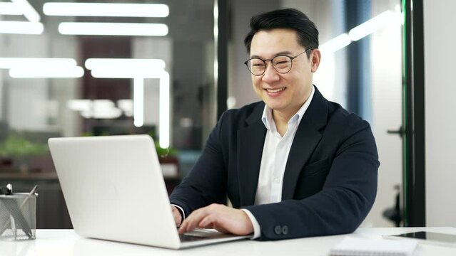 Happy asian businessman is using laptop sitting at workplace in business office. Smiling man in formal suit reads or typing message, chats online or works in computer application and looking at camera