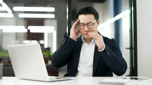 Tired asian businessman suffering from headache while working on laptop computer while sitting at desk at workplace in business office. Man in formal suit massages and rubs his temples with his hands