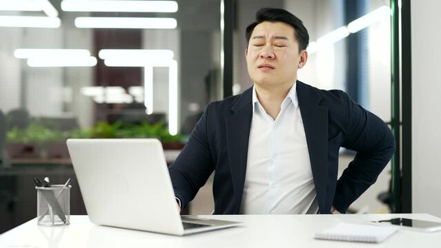 Asian businessman suffering from back pain while working on laptop sitting at workplace in business office Sick mature man in a formal suit has back muscle spasms, massages his lower back, stretches