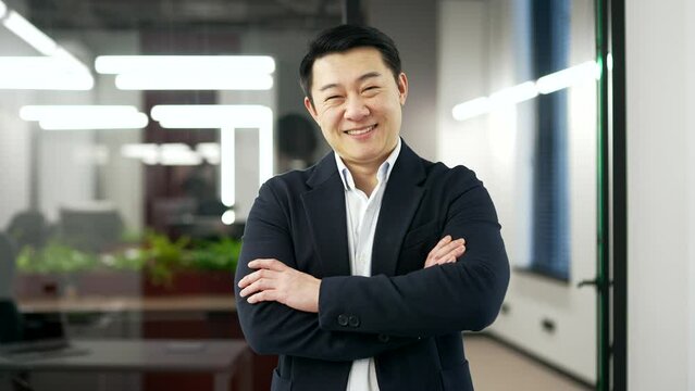 Portrait of a happy asian businessman in a formal suit standing with crossed arms in a business office. Confident smiling mature man posing looking at camera. Head shot of an entrepreneur or manager