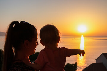 Silhouette of loving mother holding small toddler at romantic sunset in Omis Riviera, Split-Dalmatia, Croatia. Coastline South Dalmatian coast, Adriatic Sea in summer. Family travel vacation concept - obrazy, fototapety, plakaty
