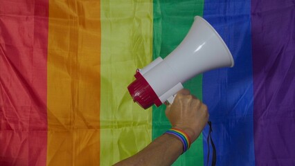 Hand with rainbow bracelet holding megaphone with LGBT flag on background. Gay rights concept