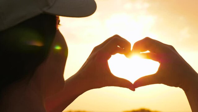 travel background. Close-up of a woman wearing a baseball cap holding her palms folded in the shape of a heart. The rays of the sun pass through the womans hands.