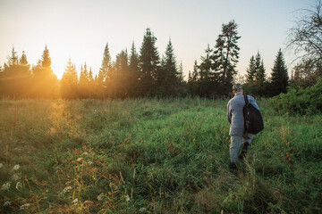 A man with a backpack is standing in a grassy meadow at sunset, surrounded by trees and natural landscape, under a colorful sky