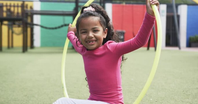 In a school playground, a young African American girl smiles with a hula hoop