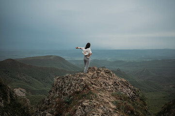 Fototapeta premium Happy woman tourist hiking up mountain enjoying nature. Landscape in cloudy gloomy windy weather. Female in white shirt with open arms outstretched in joy, enjoying travel. Freedom concept