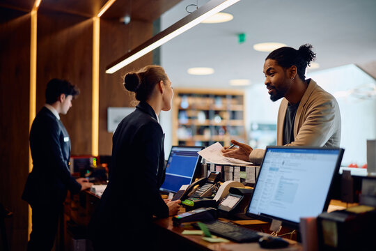 Black man talking to receptionist while signing paperwork at hotel reception desk.
