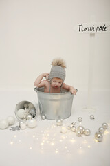 Naklejka premium a little boy sits in a metal basin on a white background in a cap. New Year's idea for photos of small children. scattered Christmas decorations. stylish children's photo zone. Merry Christmas 