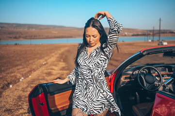 Happy smiling brunette woman driver in fashion dress posing with new red cabriolet car on beach...