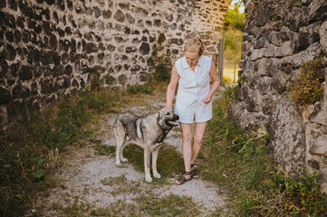 Happy smiling senior elderly mature woman tourist walking outdoors in ancient fortress in Europe hugging homeless big dog. Retired sporty active old people and animals pets concept