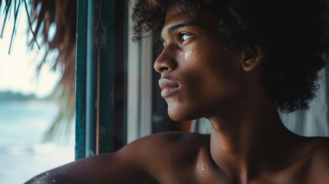 A portrait of an attractive young black teenage surfer boy in his Thailand beach hotel room looking longingly out window