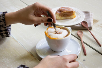 hands, with dark black nails, stirring a spoon in a cup of cappuccino with dulce de leche, on a...