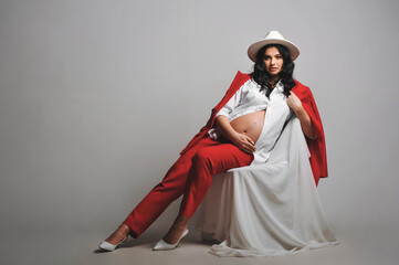 Horizontal portrait of brunette pregnant woman in white hat, shirt and red suit posing on a cyclorama. Copy space for text