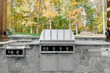 Sunny Outdoor Kitchen with Stainless Steel Grill, Side Burners Surrounded by Natural Stone and Wood Pergola Overlooking Lush Forest