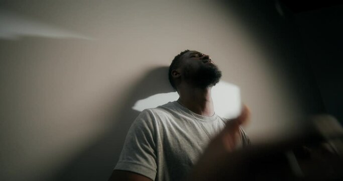 A frustrated man covers his face with trembling hands, standing near a white wall indoors