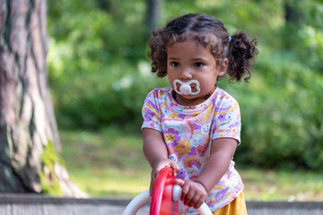A thoughtful child with a pacifier, riding a toy in the park, captures a carefree summer moment,...