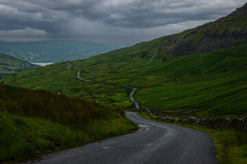Asphalt mountain road winding between green hills under dark rain clouds