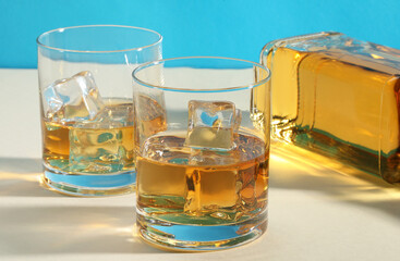 Whiskey with ice cubes in glasses and bottle on white table, closeup