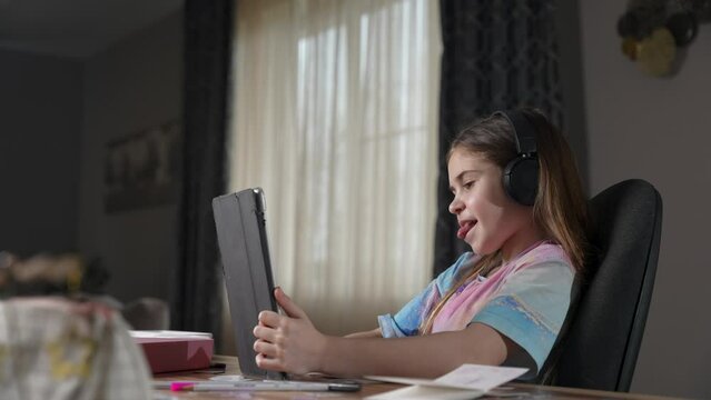Happy girl with headphones looking to tablet camera, smiling, making faces using app with masks. Child having fun, listen to music, playing, having video call. Communication online distantly
