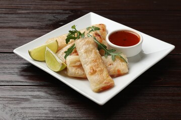 Tasty fried spring rolls, arugula, lime and sauce on wooden table, closeup