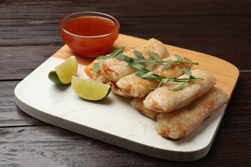 Tasty fried spring rolls, lime, arugula and sauce on wooden table, closeup