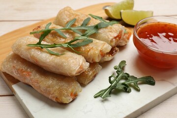 Tasty fried spring rolls, lime, arugula and sauce on light wooden table, closeup