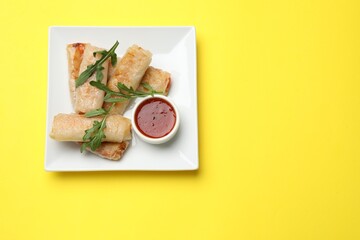 Fried spring rolls, arugula and sauce on yellow table, top view. Space for text