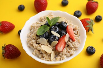 Tasty oatmeal with strawberries, blueberries and almond petals in bowl surrounded by fresh berries on yellow background