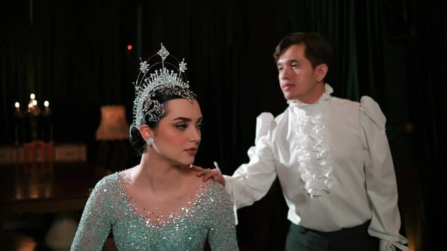 Man in vintage attire whispering to a woman in a sparkling dress and tiara against a dark background.