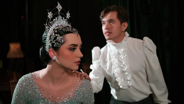 Man whispering to an elegant woman in a sparkling tiara and costume, depicting secrecy or gossip in a vintage setting.
