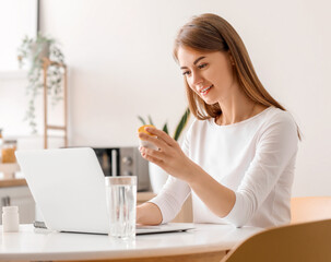 Beautiful woman with bottle of vitamin supplements using laptop in kitchen