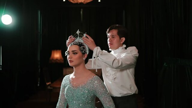 Elegant woman in a sparkling gown with a crown being adjusted by a man on a dark stage with dramatic lighting.