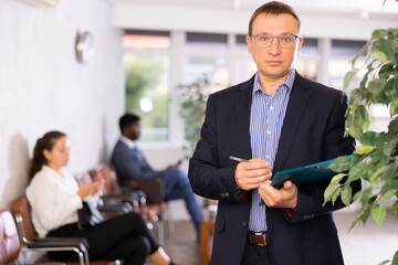 Dispassionate secretary man in glasses stands at entrance to office reception with folder and list...